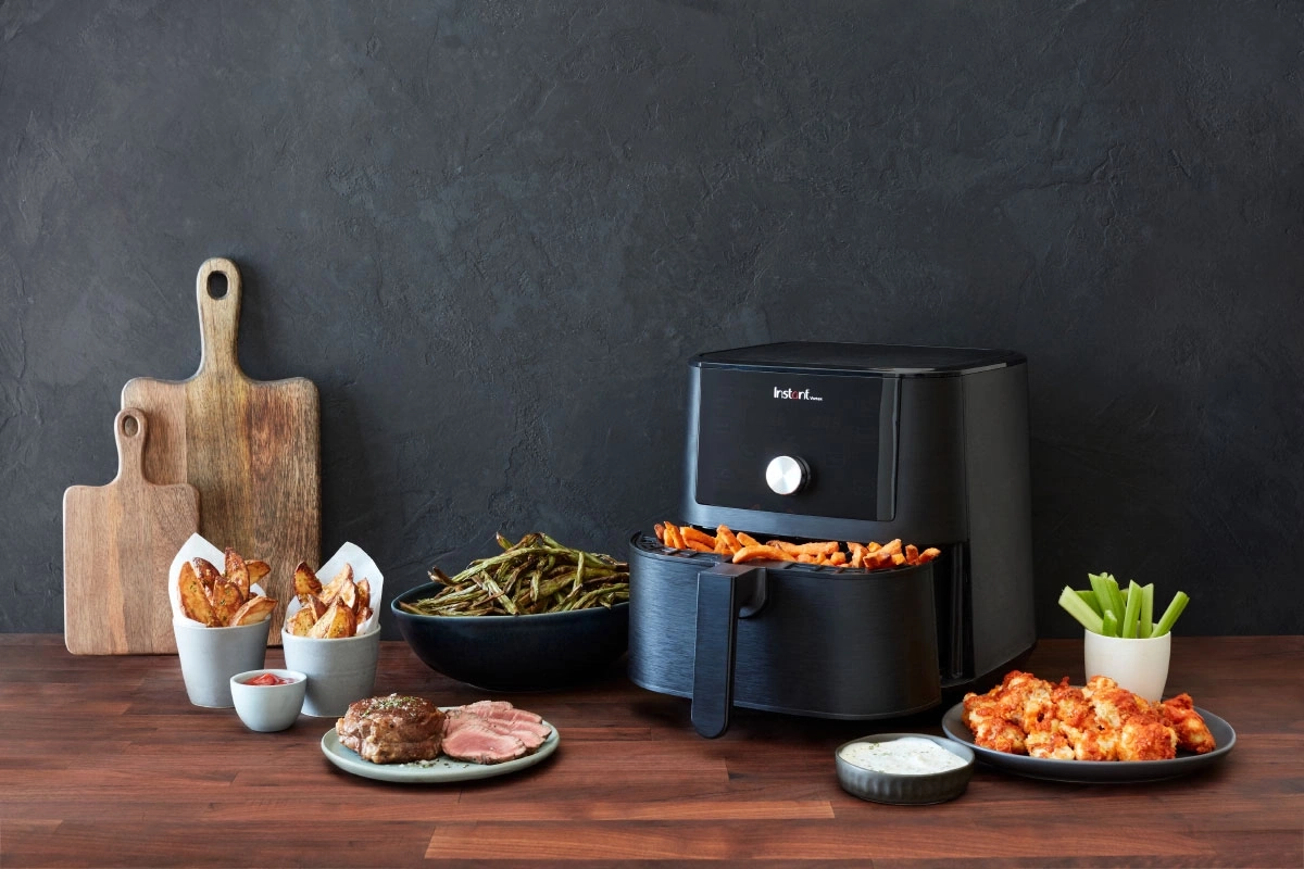 Schwarzer Airfryer mit Essen, Holzbrettern und verschiedenen Gerichten auf einem Holztisch.